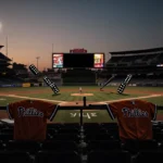 Empty baseball stadium seats cast long shadows with Phillies jerseys draped over railing and bat leaning nearby