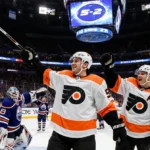 Denver Barkey raises his stick proudly with Flyers teammates cheering and scoreboard showing 5-2 in the electric arena