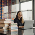 Woman smiles while working on laptop at desk with Ukrainian textiles and language books