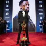 Ryan Coogler standing on the red carpet with a gold statuette beside him and a large screen showing Sinners behind him