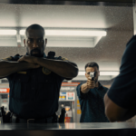 Security guard shielding himself with arms up against a gas station counter while a shadowed man holds a gun in background.