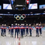 Speed skaters gather on shiny ice with arena lights reflecting and Olympic rings on scoreboard as Milwaukee fans cheer