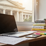 Student studying at desk with laptop and books and Indian flag visible through window