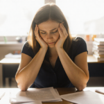 Teacher sits at desk with head in hands and papers while warm window light highlights her contemplative face concerned mental