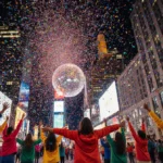 Volunteers raise arms with confetti bursting above and Times Square ball glowing in distance