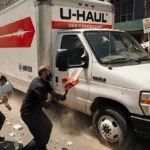 U-Haul truck drives through protesters with angry crowd surrounding vehicle and signs waving