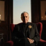 Valentino Garavani sits in crimson armchair with golden brooch glimmering and tears reflecting warm candlelight in opulent Ro