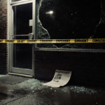 Police tape wraps around a broken storefront window of convenience store at nighttime with newspaper clipping on ground