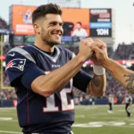 Zac Efron high-fiving Tom Brady at Patriots game with fans cheering and scoreboard glowing behind