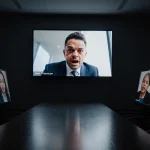 A distorted face interrupts a Zoom meeting with shocked board members visible on screen and darkness filling the empty room